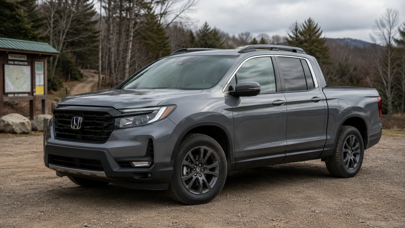 A gray 2026 Honda Ridgeline pickup truck parked at a trailhead in overcast daylight, shot candidly at a three-quarter angle.
