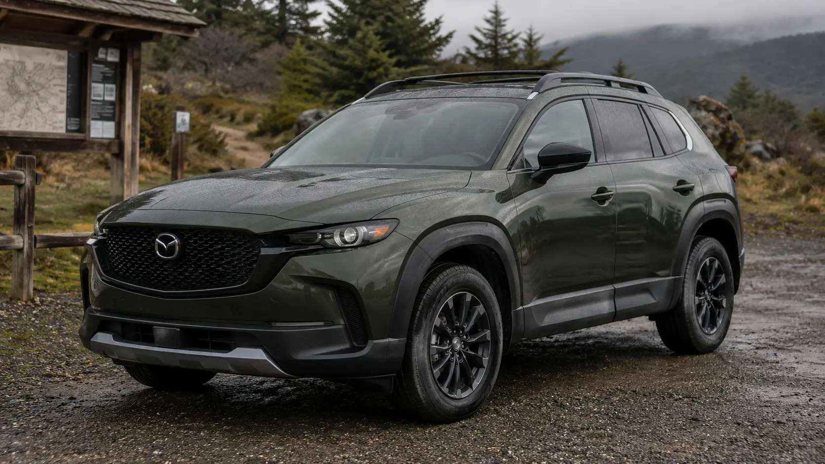 A dark green 2026 Mazda CX-50 crossover parked at a trailhead in overcast daylight, shot candidly at a three-quarter angle.