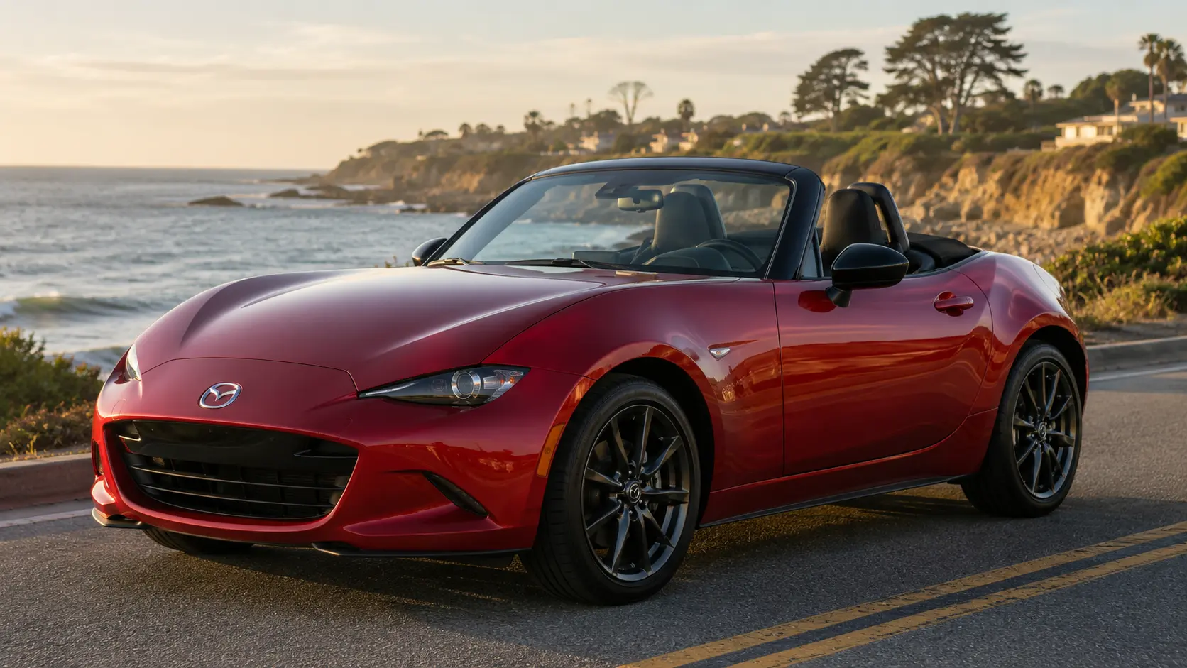 A red 2026 Mazda MX-5 Miata convertible with the top down, parked along a coastal road in late-afternoon light, shot candidly at a three-quarter angle.